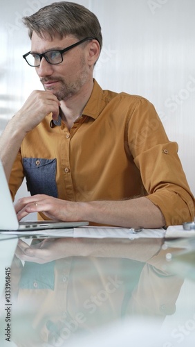 Obraz na płótnie Focused businessman in yellow casual shirt is using laptop and thinking with hand on chin, working on a project in a modern office, vertical portrait. Business people concept w salonie