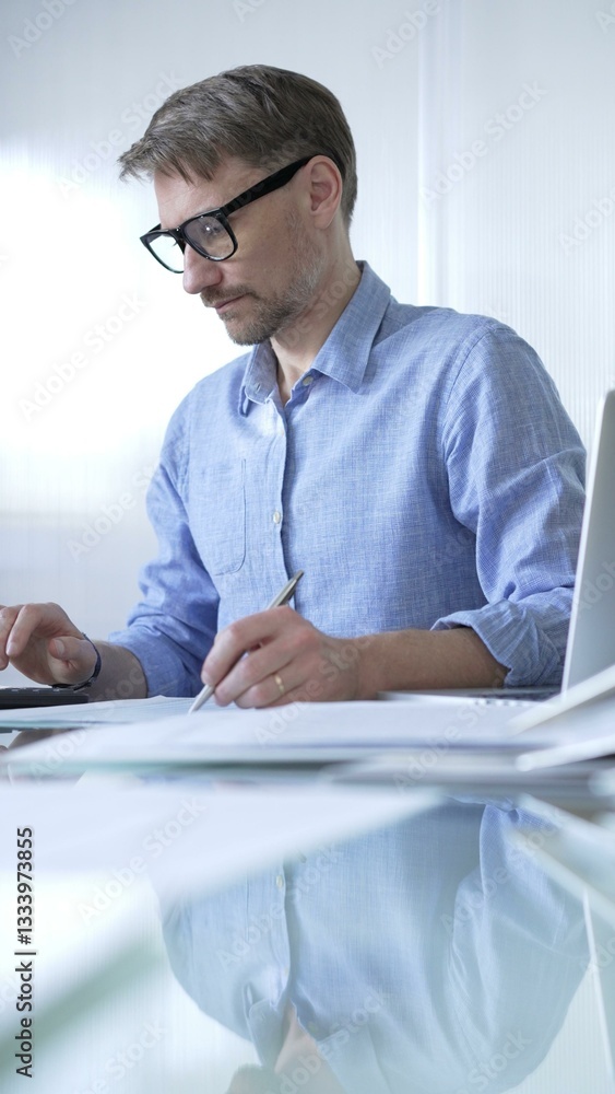 Businessman or accountant, wearing blue casual shirt and glasses, is working on tax calculations, note taking, and financial reports in the office. Audit and taxes in business