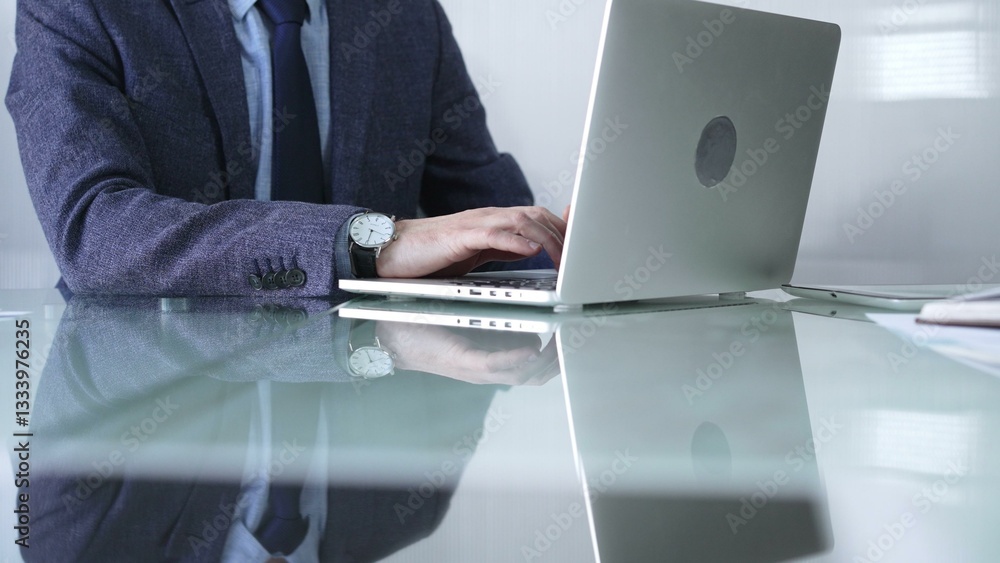 Businessman in blue jacket is focused on typing on laptop at glass desk in modern office, showcasing productivity and concentration, closeup view. Business people concept