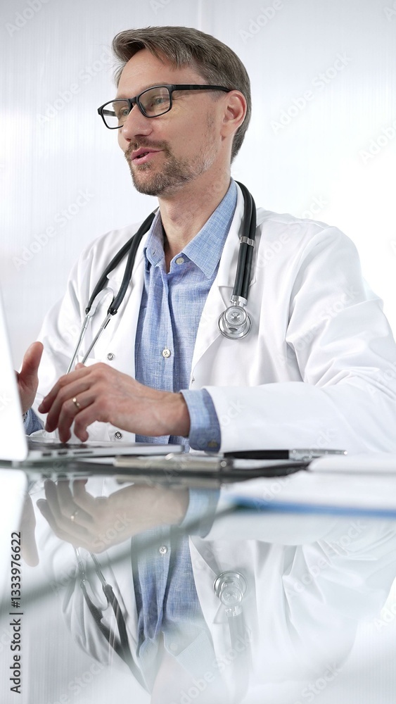 Doctor man, wearing a stethoscope, blue shirt and a lab coat is gesturing with hands overs a glass desk with laptop in clinic office, vertical portrait view. Medicine and science Doctor man, wearing a stethoscope, blue shirt and a lab coat is gesturing with hands overs a glass desk with laptop in clinic office, vertical portrait view. Medicine and science