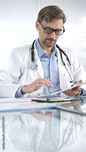 Obraz na płótnie Doctor man in blue shirt under a white coat, glasses and stethoscope is using digital tablet in modern healthcare setting, interacting with patient data, vertical portrait view. Medicine and science w salonie