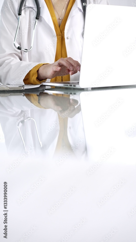 Unknown doctor woman, wearing a stethoscope and yellow blouse under a lab coat, is using technology in healthcare. Close-up of hands typing on a laptop on a glass desk, vertical view. Medical concept