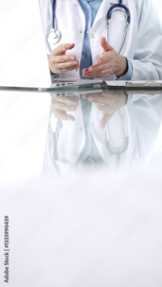 Unknown doctor woman, wearing a stethoscope, blue blouse under a lab coat, is gesturing with hands overs a glass desk with a laptop in clinic office, close up vertical view. Medicine and science