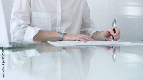 Obraz na płótnie Businessman in white casual shirt is signing a contract at a sleek glass desk in a modern office, showcasing professionalism and focus in his work environment. Business people concept w salonie