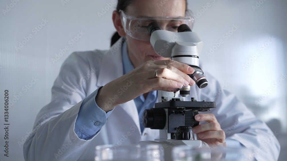 Woman scientist wearing lab coat and protective glasses is using microscope in modern laboratory setup. Low key lighting photo. Science and Medicine Woman scientist wearing lab coat and protective glasses is using microscope in modern laboratory setup. Low key lighting photo. Science and Medicine