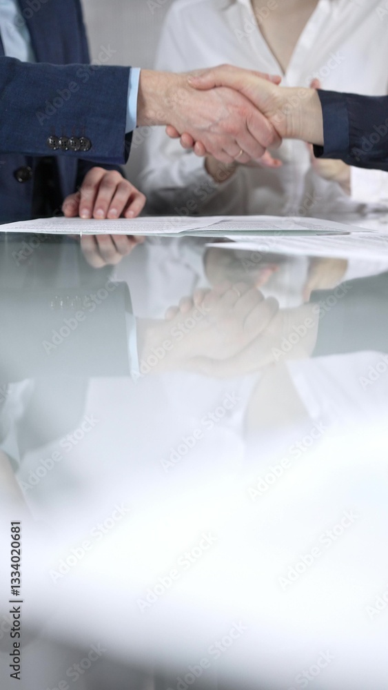 Business partners shaking hands after signing contract at glass table in office meeting, making a deal and closing agreement, vertical close up of handshake. Copy space area