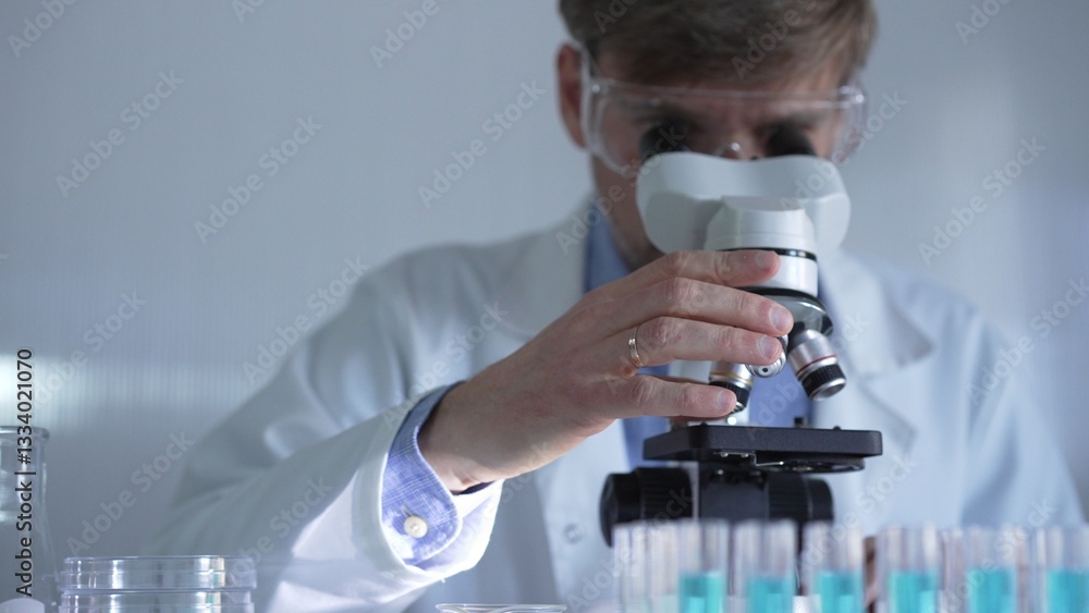 A man scientist, wearing a lab coat, mask, and safety glasses, is using a microscope surrounded by test tubes in laboratory setting. Medicine and science concept