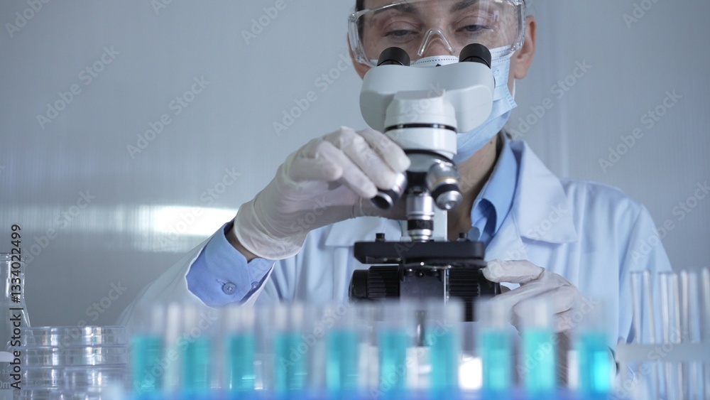 A scientist woman, wearing a lab coat, mask, white gloves and safety glasses, is adjusting a microscope surrounded by test tubes in laboratory