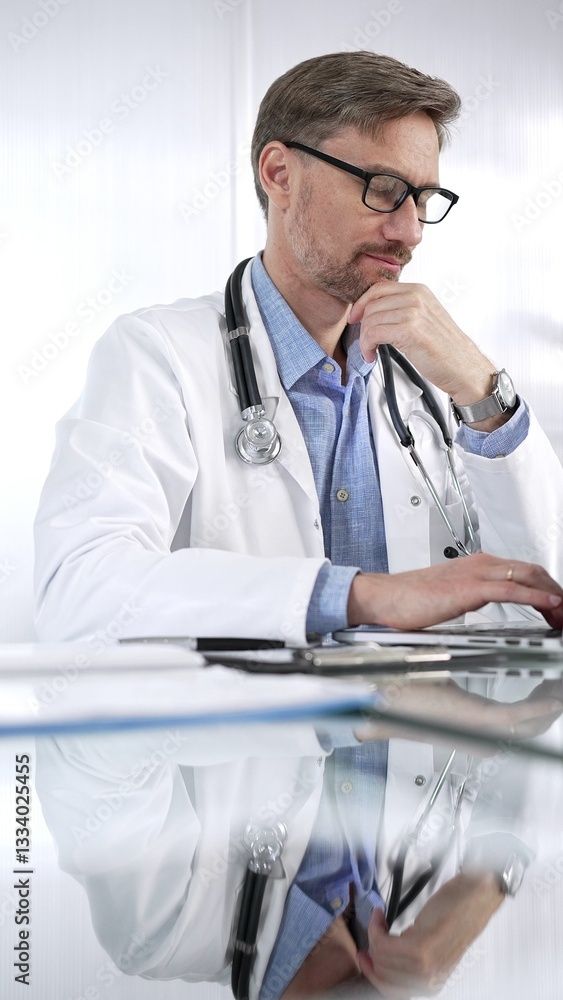 Doctor man in a blue shirt under a medical coat is thinking while using technology in medicine. Portrait of physician typing on a laptop on a glass desk in clinic, vertical view. Medicine and science