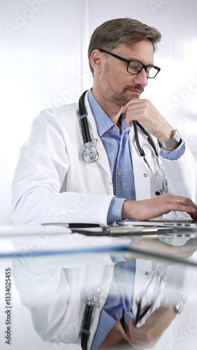 Obraz na płótnie Doctor man in a blue shirt under a medical coat is thinking while using technology in medicine. Portrait of physician typing on a laptop on a glass desk in clinic, vertical view. Medicine and science w salonie