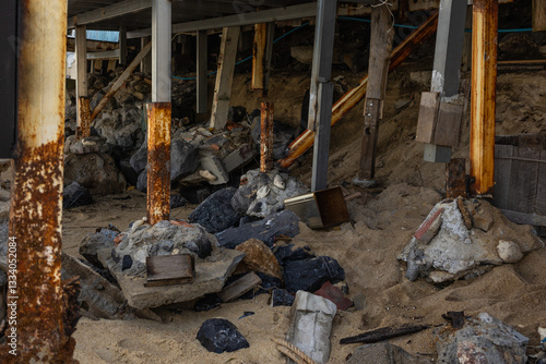Rusty metal beams and broken concrete under a damaged structure. Debris, sand, and black rocks scattered, showing destruction and decay.