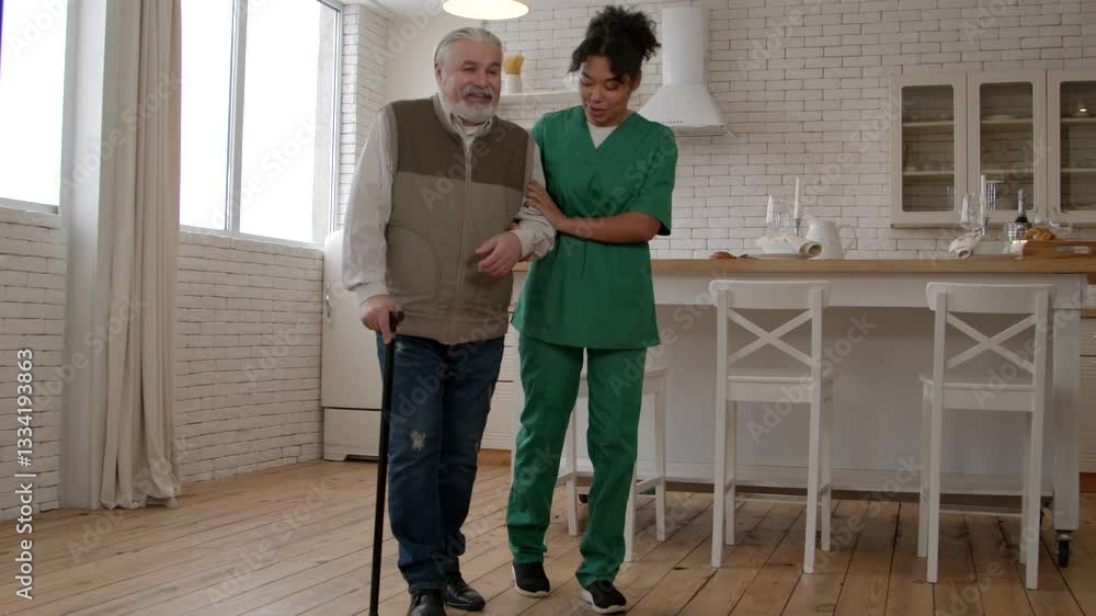 Positive handsome retired bearded man with help of lovely African American female nurse in uniform walking along nursing home using walking stick, friendly communicating and sharing. Positive handsome retired bearded man with help of lovely African American female nurse in uniform walking along nursing home using walking stick, friendly communicating and sharing.