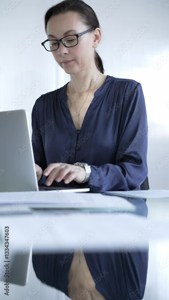 Focused businesswoman with glasses typing on laptop at modern glass desk in well lit office. Business people concept Focused businesswoman with glasses typing on laptop at modern glass desk in well lit office. Business people concept
