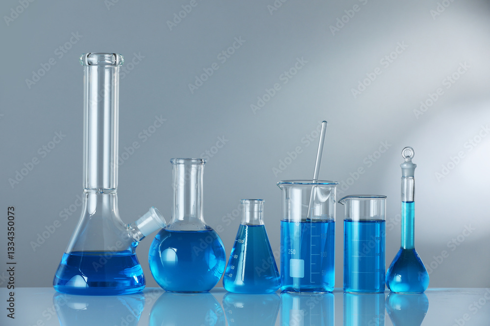 Laboratory glassware with blue liquid on mirror surface against grey background. Space for text Laboratory glassware with blue liquid on mirror surface against grey background. Space for text