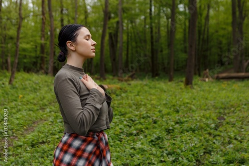 Young woman standing in verdant forest, palms resting on chest, experiencing peaceful moment while breathing deeply and connecting with surrounding woodland environment
