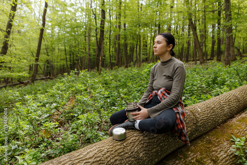 Female hiker sipping warm beverage from thermos, sitting on fallen tree trunk, surrounded by verdant forest foliage, experiencing peaceful wilderness moment