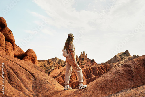 Girl traveler with backpack stands on the edge of cliff and looks at beautiful view of Skazka canyon in Kyrgyzstan.