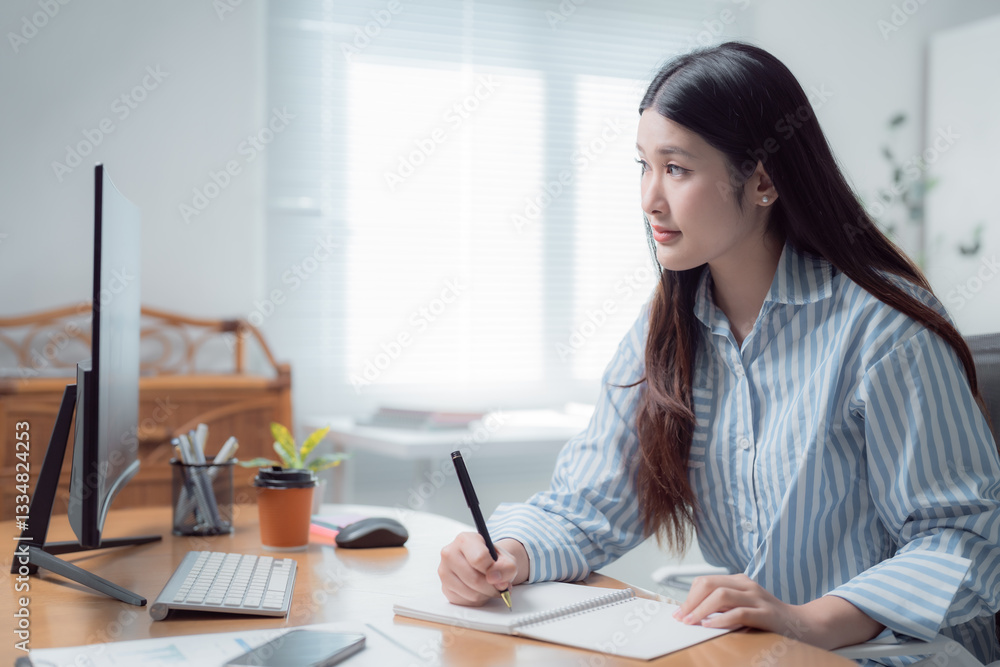 Young Asian businesswoman taking notes in her notebook while focused on the computer screen, working efficiently from her cozy home office during the day, enjoying natural light Young Asian businesswoman taking notes in her notebook while focused on the computer screen, working efficiently from her cozy home office during the day, enjoying natural light