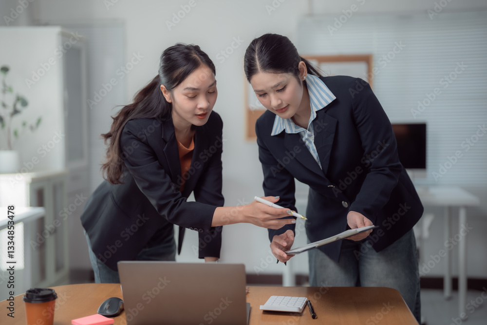 Two young businesswomen are working together in modern office, analyzing financial data using laptop and documents, pointing at charts and discussing project strategy Two young businesswomen are working together in modern office, analyzing financial data using laptop and documents, pointing at charts and discussing project strategy
