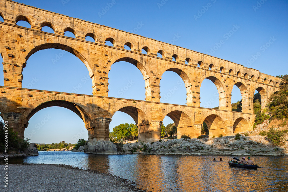 Fototapeta premium Pont du Gard, ancient Roman aqueduct bridge, Provence late afternoon golden light on arches mirroring in river, Gorges du Gardon, natural landscape designated Biosphere Reserve by UNESCO, France