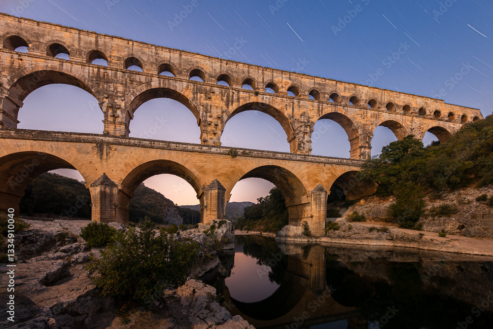 Fototapeta premium Pont du Gard, ancient Roman aqueduct bridge, colorful distant sunrise, under Provence starry night sky, Gorges du Gardon, natural landscape designated Biosphere Reserve by UNESCO, France