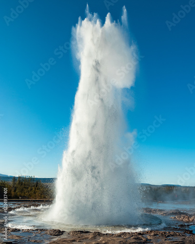 Strokkur geyser in Iceland erupts, shooting boiling water high into the air. A popular attraction in the Golden Circle, showcasing Iceland’s geothermal activity on a clear day with a blue sky. 