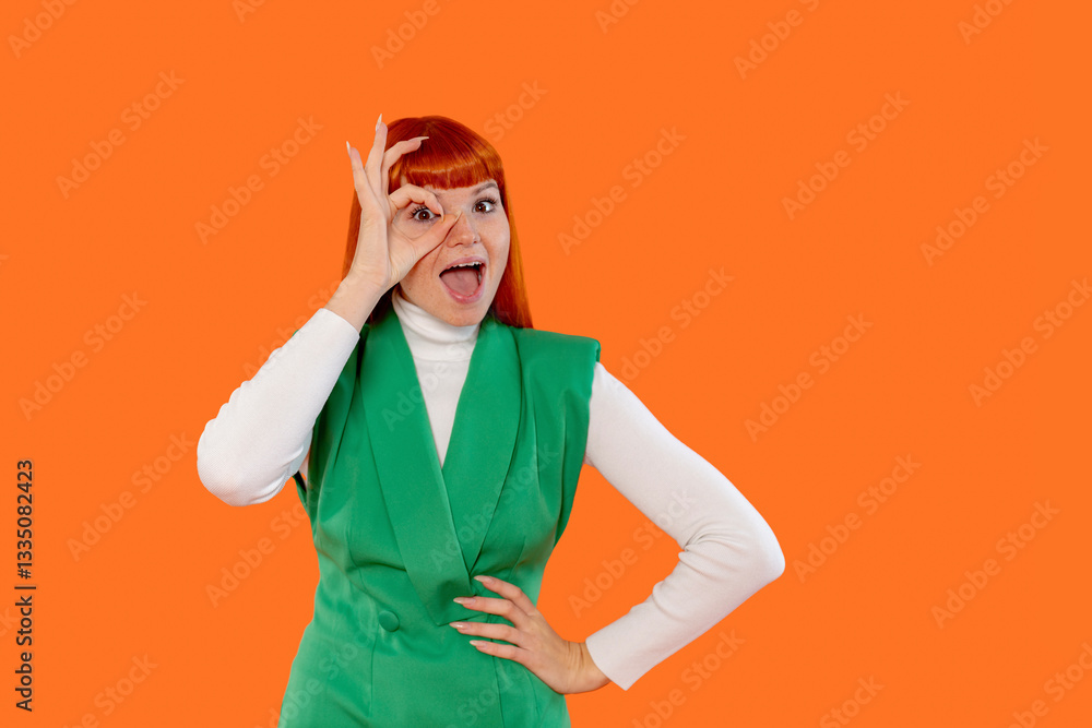 Young woman displaying playful expression with hand gesture against solid orange background while wearing green outfit Young woman displaying playful expression with hand gesture against solid orange background while wearing green outfit