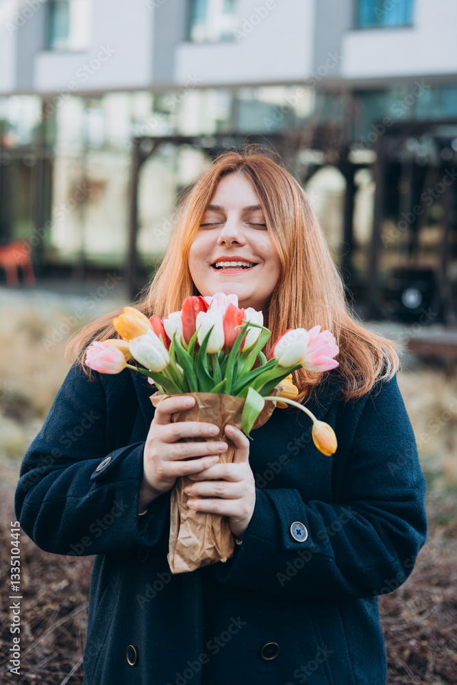 Beautiful young woman with spring tulips flowers bouquet walking at city street. Happy girl posing outdoors. Spring portrait of pretty female on urban background. High quality photo.