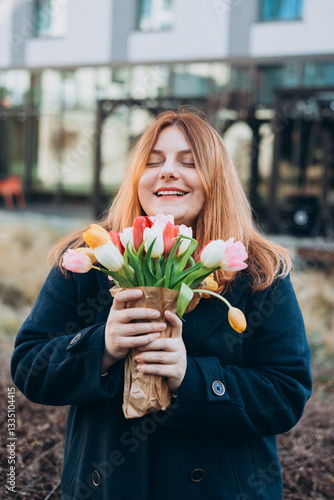 Obraz na płótnie Beautiful young woman with spring tulips flowers bouquet walking at city street. Happy girl posing outdoors. Spring portrait of pretty female on urban background. High quality photo. w salonie