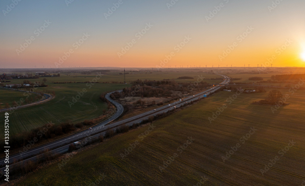 Fototapeta premium Aerial view of a highway cutting through vast rural fields under a golden sunset. The warm light casts long shadows, highlighting the peaceful countryside and the contrast between nature and infrastru