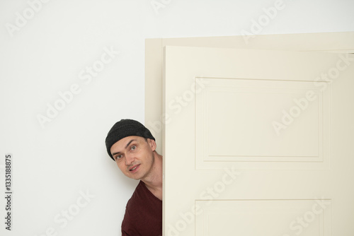 A man wearing a beanie leans playfully around an open door in a bright, minimalistic space. His curious expression adds life to the simple decor