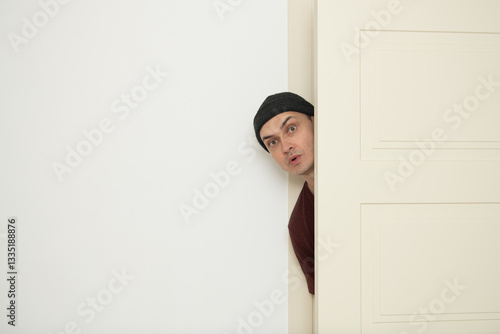 A person with a beanie is curiously peeking around an open door, exhibiting a surprised expression. The surrounding space is minimalist with white walls