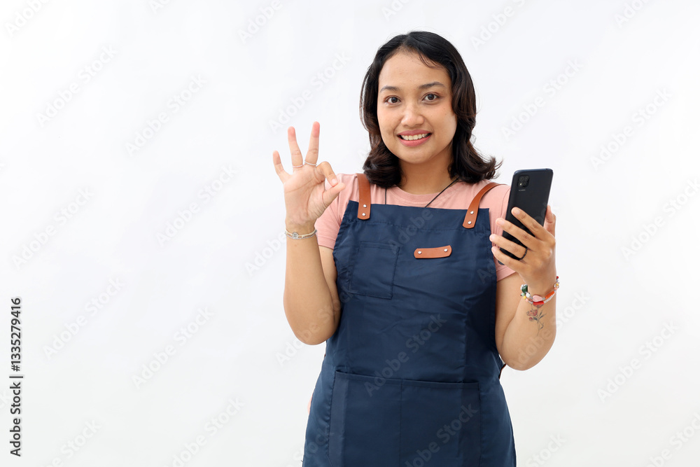 Smiling asian barista woman in apron holding smartphone and showing ok sign gesture on white background. Smiling asian barista woman in apron holding smartphone and showing ok sign gesture on white background.