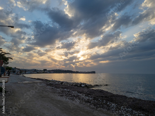 Sun setting over a calm ocean reflecting golden light, casting shadows across the cloudy sky.