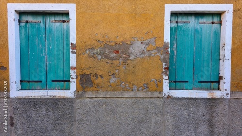 A weathered yellow wall with peeling plaster features two closed turquoise wooden shutters framed in white. The aged texture and faded colors create a rustic, charming aesthetic.