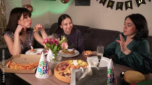 Friends gather at home, at the festive table, celebrating a fun and unforgettable birthday. There are pizzas, cakes and various snacks on the table, as well as birthday caps.