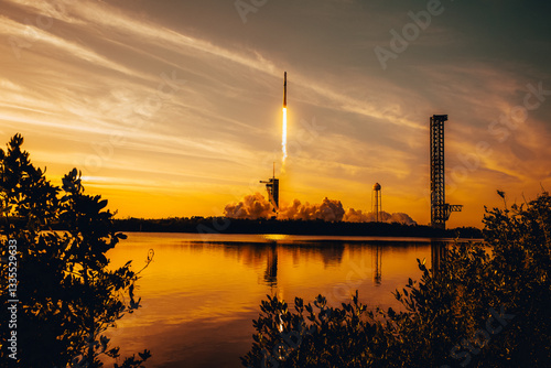 Fototapeta Naklejka Na Ścianę i Meble -  Rocket launch at sunset with fiery trail over water, reflected in golden hues. SpaceX Falcon rocket blasting off. Exploration, technology and space concept. Elements of this image furnished by NASA