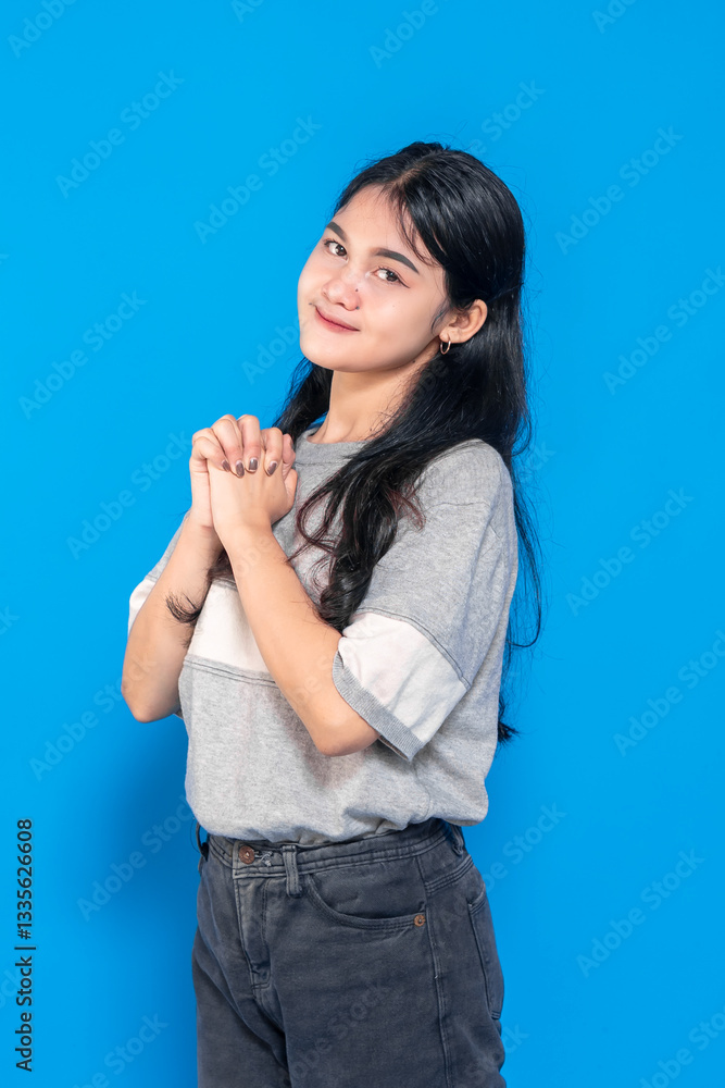 Young Asian woman with a hopeful expression, hands clasped together, smiling gently. She looks grateful and excited, expressing optimism against a vibrant blue background. Young Asian woman with a hopeful expression, hands clasped together, smiling gently. She looks grateful and excited, expressing optimism against a vibrant blue background.