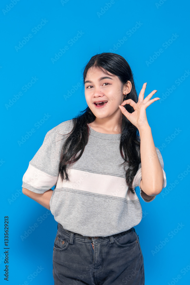 Young Asian woman smiling and making an OK hand gesture. She has long black hair, wears a casual gray t-shirt, and stands against a solid blue background, expressing positivity. Young Asian woman smiling and making an OK hand gesture. She has long black hair, wears a casual gray t-shirt, and stands against a solid blue background, expressing positivity.