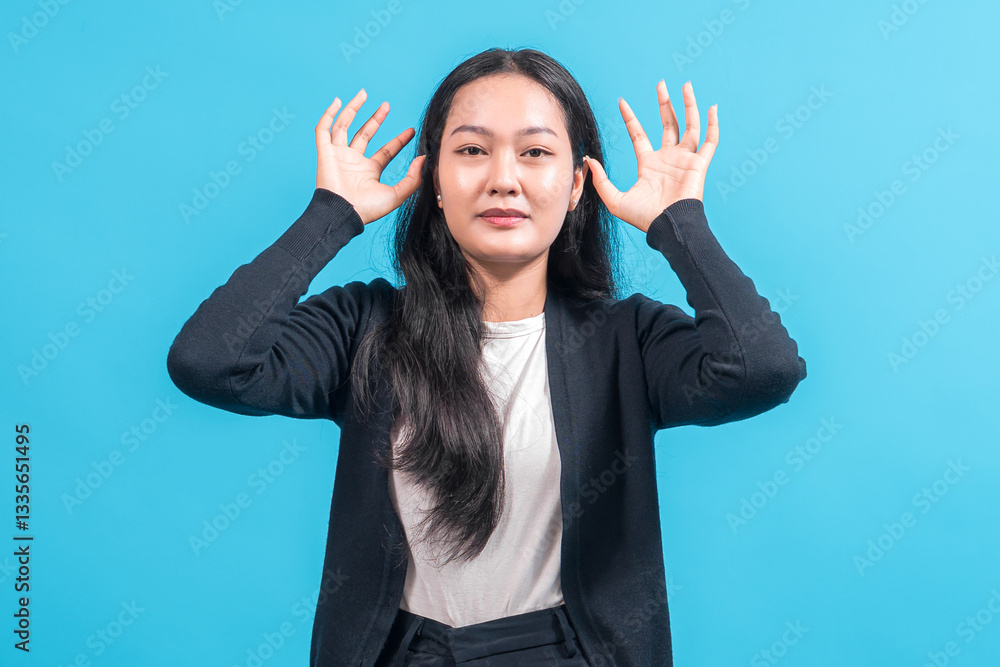 Asian woman with long black hair wearing a black cardigan and white shirt, standing against a blue background, raising both hands near her ears while making a playful gesture. Asian woman with long black hair wearing a black cardigan and white shirt, standing against a blue background, raising both hands near her ears while making a playful gesture.
