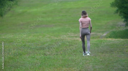 Rear view of unrecognizable female fitness instructor walking across field in short grass during summer sports training. Slim sportsperson dressed in sportswear - t-shirt, yoga pants and sports shoe
