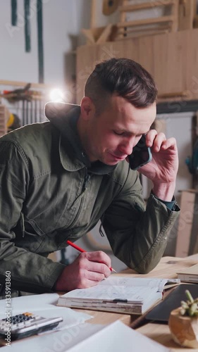 Carpenter talking to a furniture customer on the phone in his own workshop. A carpenter, a small business owner in a carpentry shop, talks to a customer on the phone.