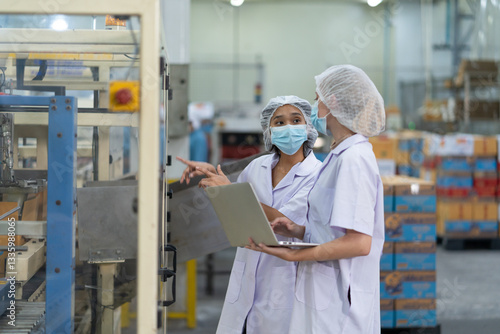 Two female workers wearing protective clothing and face masks conduct a quality control inspection in food processing facility. One holds laptop at the production machine.