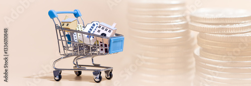 miniature shopping cart filled with small houses beside stacks of coins, symbolizing real estate investment, home buying, and property market trends.
