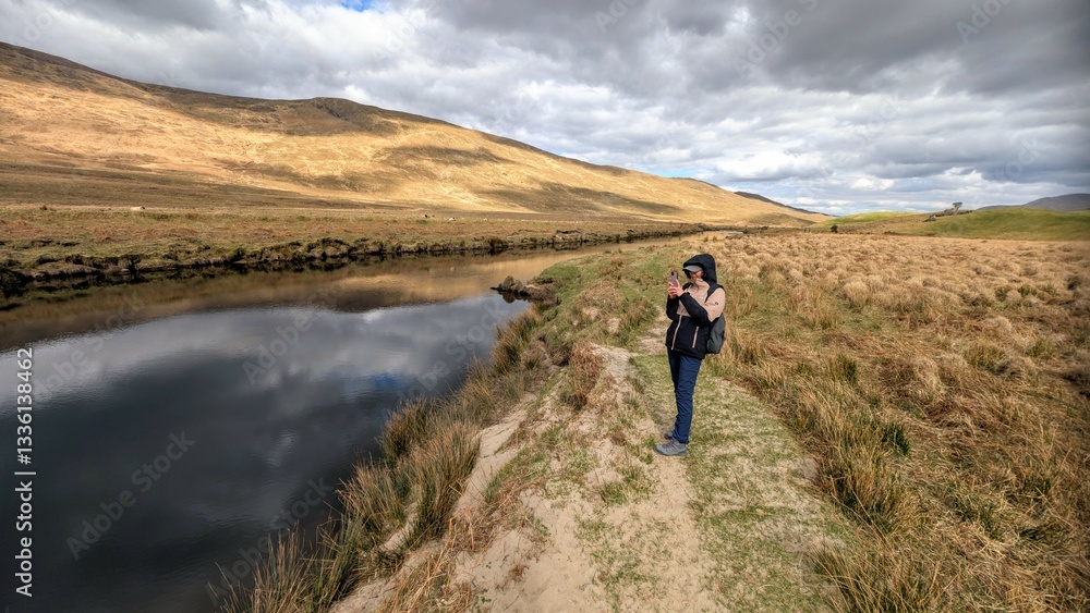 Beautiful woman backpacker in hooded jacket hiking at river Erriff near Aesleagh, county Mayo, Ireland, Irish landscape, mountains and nature background