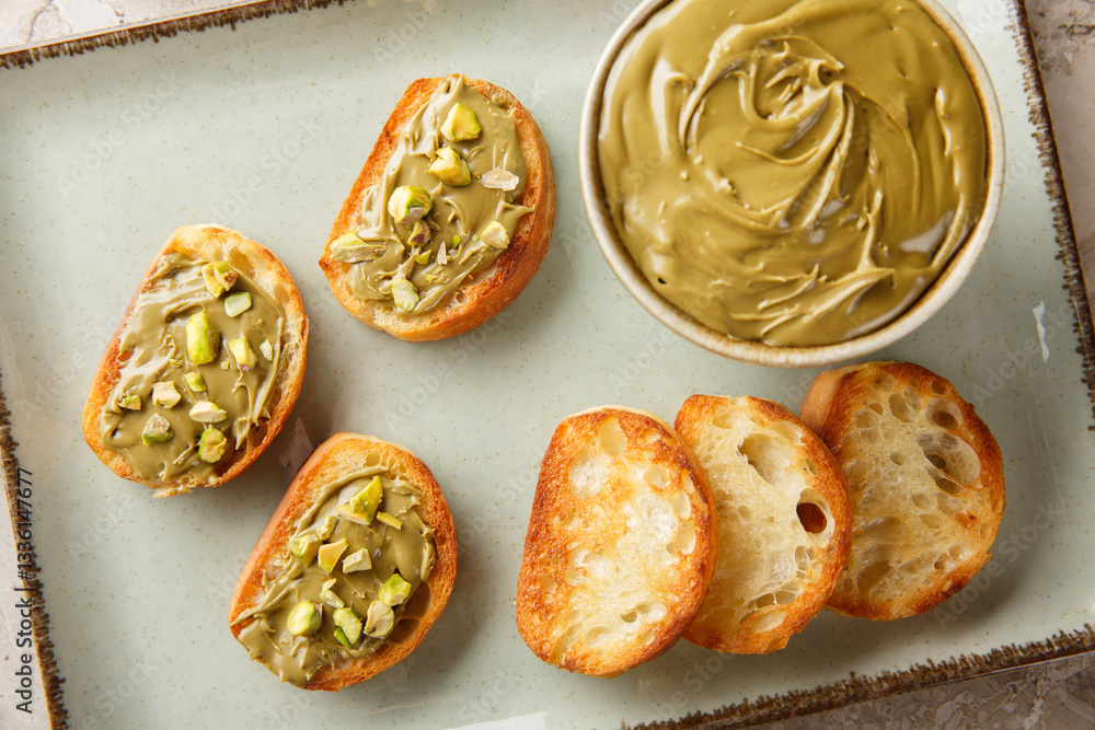 Pistachio spread in bowl and toasted bread close-up on plate on table. Horizontal top view from above
