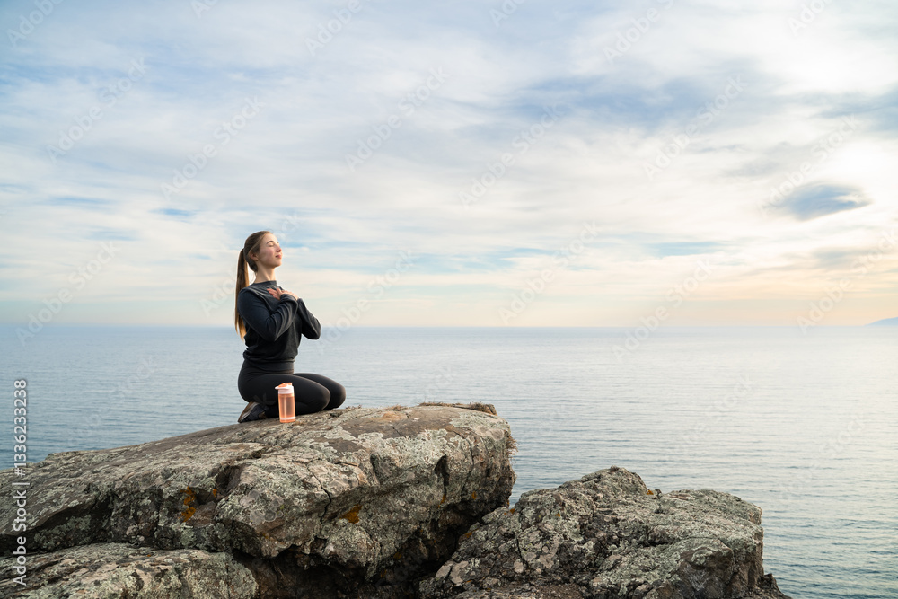 A young woman practicing gratitude meditation on a rocky cliff overlooking the sea ocean. Mindfulness, relaxation, inner peace, calm, mental health, spiritual, healing, outdoor wellness, self-care. A young woman practicing gratitude meditation on a rocky cliff overlooking the sea ocean. Mindfulness, relaxation, inner peace, calm, mental health, spiritual, healing, outdoor wellness, self-care.