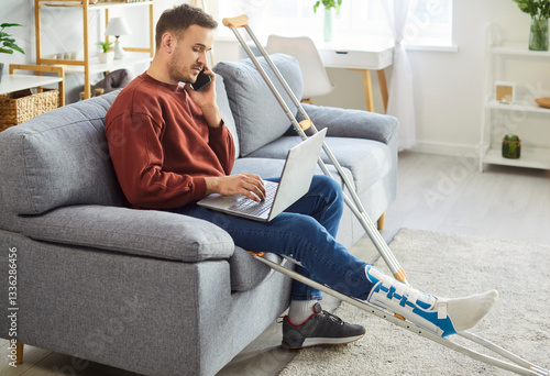 Obraz na płótnie Young man with broken leg working on laptop at home talking on mobile phone sitting on sofa with crutches after accident