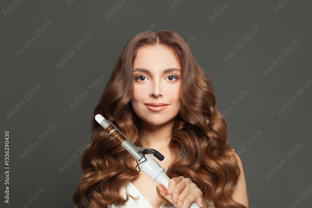Obraz premium Pretty brown haired woman with shiny long curly hairstyle showing curling iron. Hair treatment, hair care and styling