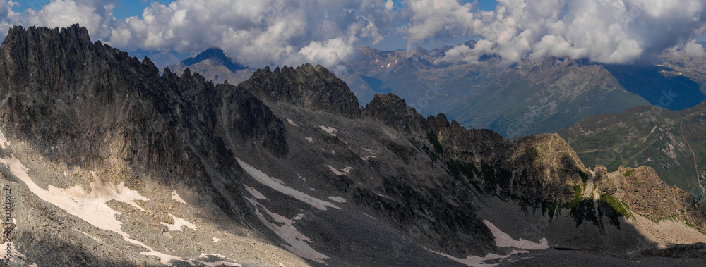 Obraz premium A wide panoramic shot capturing the rugged mountain range with patches of snow and rocky terrain, under a partly cloudy sky.